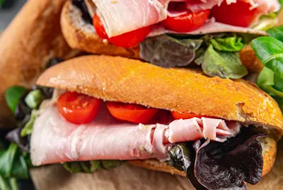 Two sandwiches on crusty bread filled with slices of ham, cherry tomato halves, and leafy greens, placed on a piece of parchment paper.