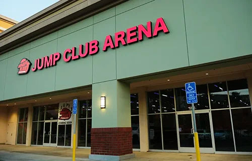 The exterior of a building with large glass windows and a red sign reading "Jump Club Arena." Two handicap parking signs are visible near the entrance.