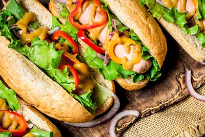 Three hot dogs in whole wheat buns topped with leafy lettuce, sliced red bell peppers, red onion, and a drizzle of mustard, arranged on a rustic wooden surface.