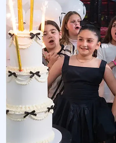 A group of children dressed for a party stand around a tall white tiered cake decorated with small black bows and sparklers, smiling and looking excited.