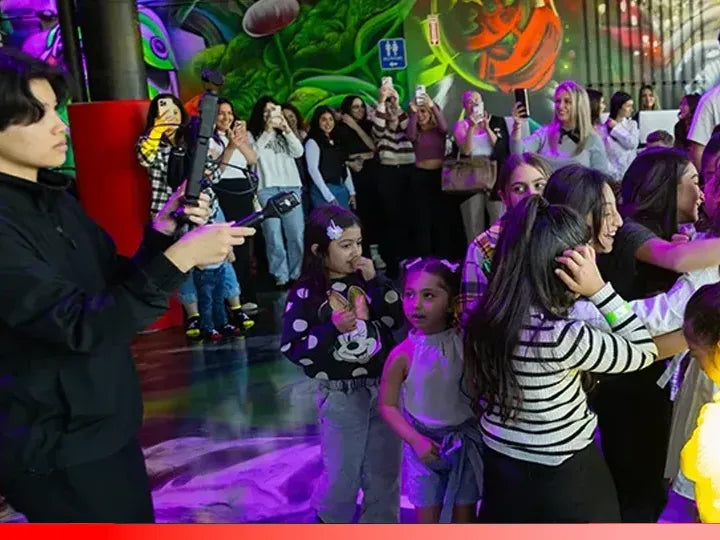 A group of children and adults, some holding phones and taking photos, gather in a colorful indoor space with vibrant wall art. The scene appears lively and festive, with a mix of excitement and engagement.