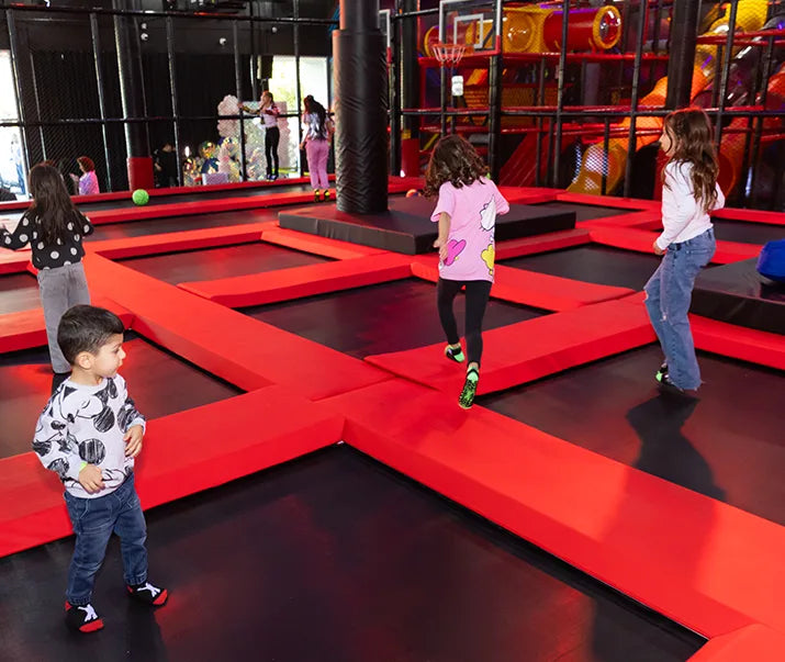 Several children play and jump on red and black trampolines indoors. In the background, there's a colorful playground structure and a basketball hoop. The atmosphere is lively and energetic.