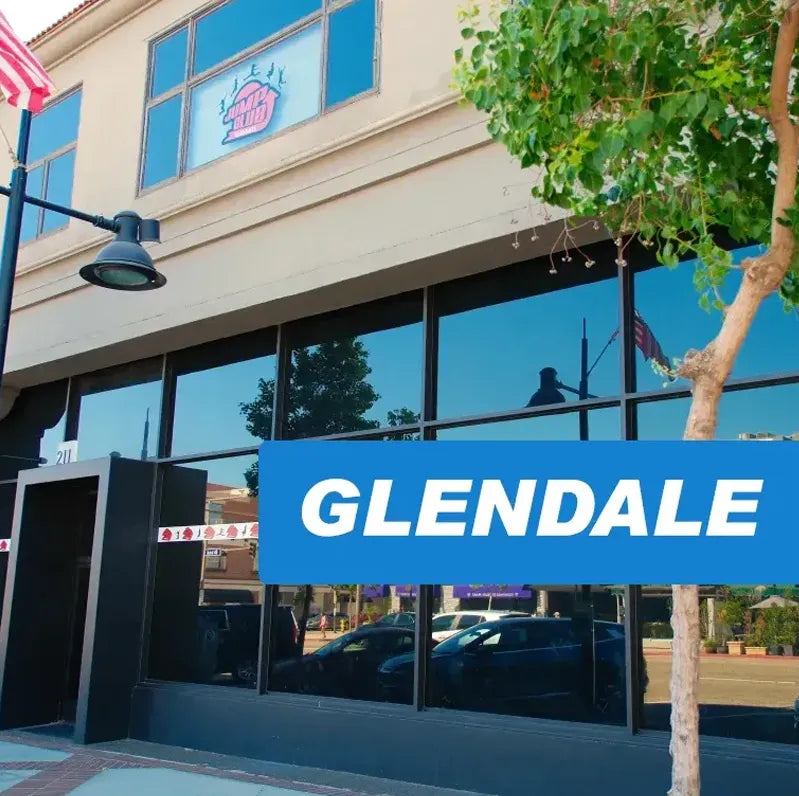 A building with large reflective windows and a sign overlay that reads "GLENDALE." An American flag and a tree are visible near the sidewalk in front.