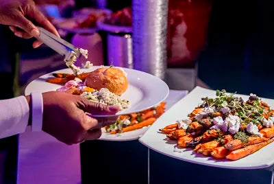 A person serves roasted carrots topped with cheese and herbs onto a plate that already holds a bread roll and a salad at a buffet.