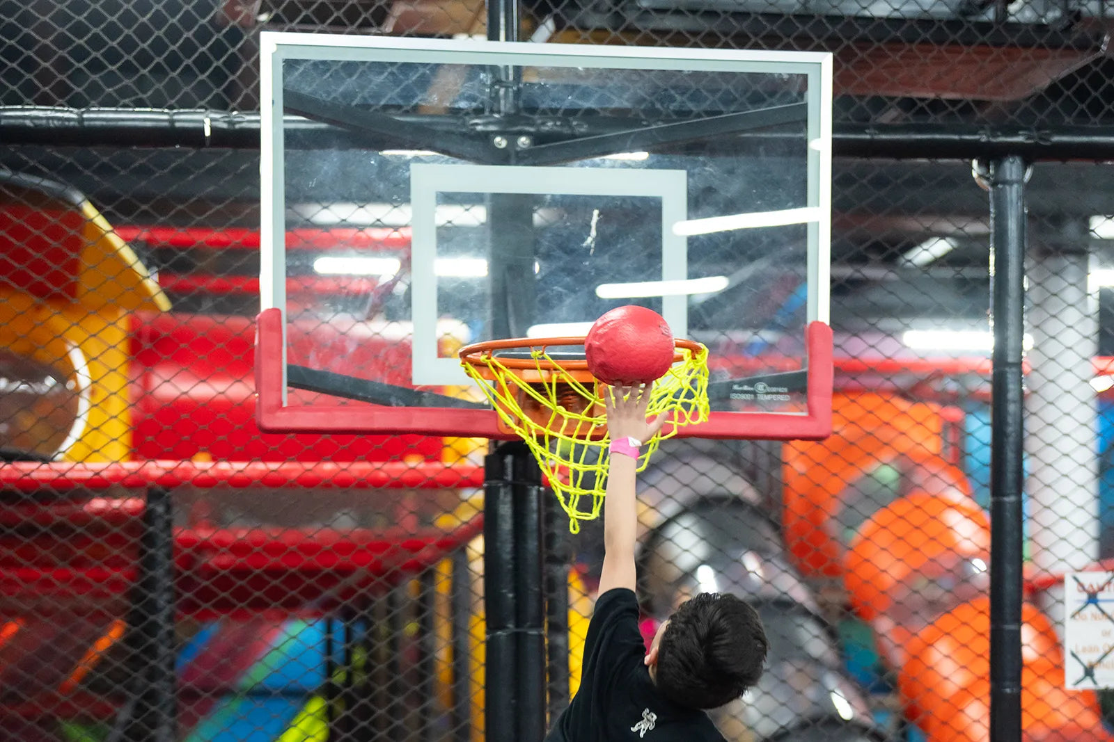 A child is jumping to dunk a red basketball into a yellow net on an indoor basketball hoop. Brightly colored playground equipment and a black chain-link fence are visible in the background.