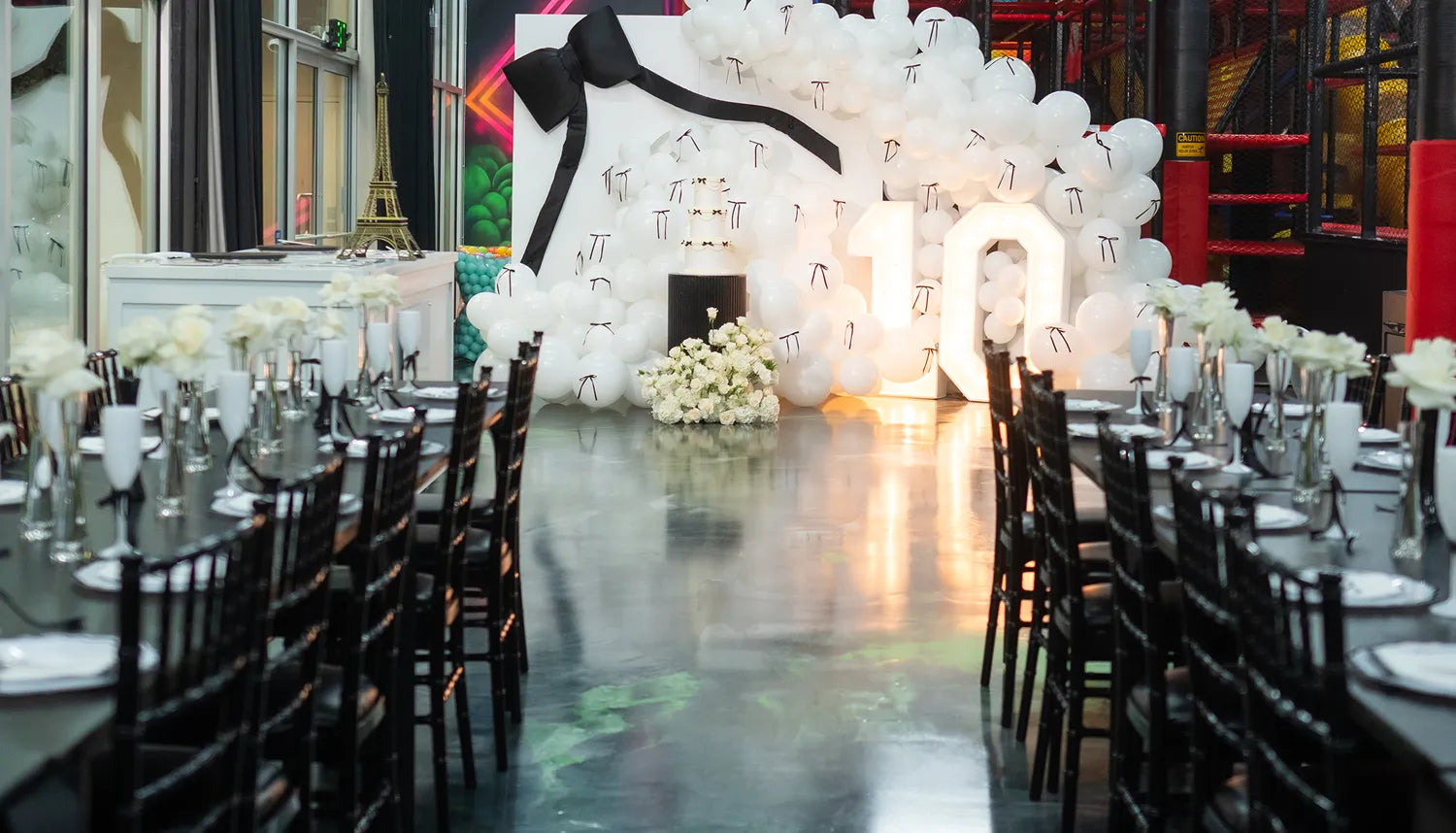 A decorated party venue with long dining tables, black chairs, white plates, and flower centerpieces. At the end, a balloon backdrop with a large "10" and lots of white balloons, some with black ribbons, is displayed.