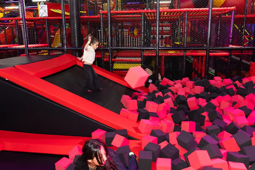 Children play in an indoor trampoline park, climbing and jumping into a foam pit filled with red and black foam cubes, surrounded by colorful play structures and safety nets.