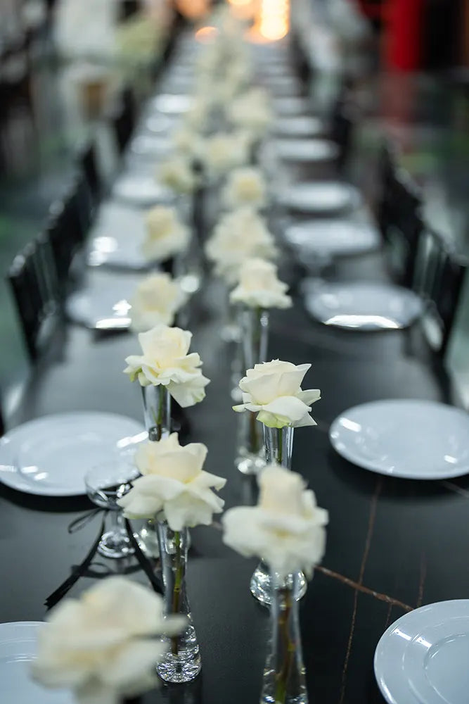 A long dining table is set with white plates and clear vases, each holding a single white rose, creating an elegant and symmetrical arrangement.