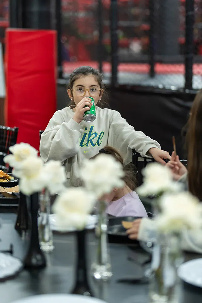A young girl wearing glasses and a light-colored Nike sweatshirt drinks from a can at a table decorated with white flowers in vases. She looks at the camera while seated among others.