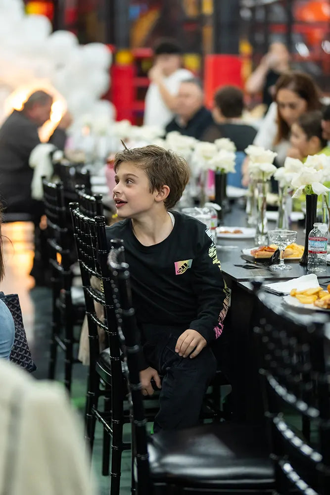 A young boy sits sideways on a black chair at a decorated table with white flowers, snacks, and drinks. Other people are seated around him in a festive, indoor setting.