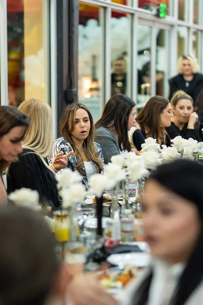 A group of women sit at a long, elegant table decorated with white flowers, engaged in conversation during a social gathering or event.