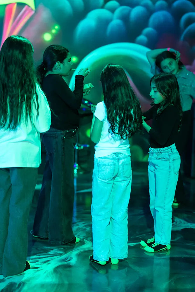 A group of children stand together in a colorful, glowing environment, possibly at an indoor event or party, talking and interacting under bright neon lights.