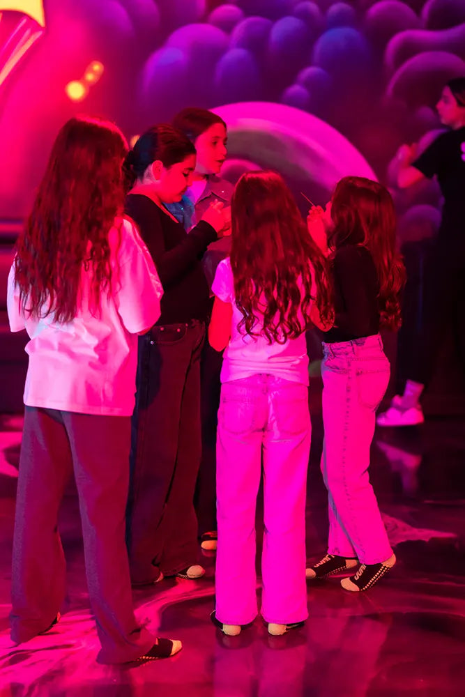 A group of five girls stand in a circle chatting under pink and purple lights at what appears to be a party or event, with another person visible in the background.