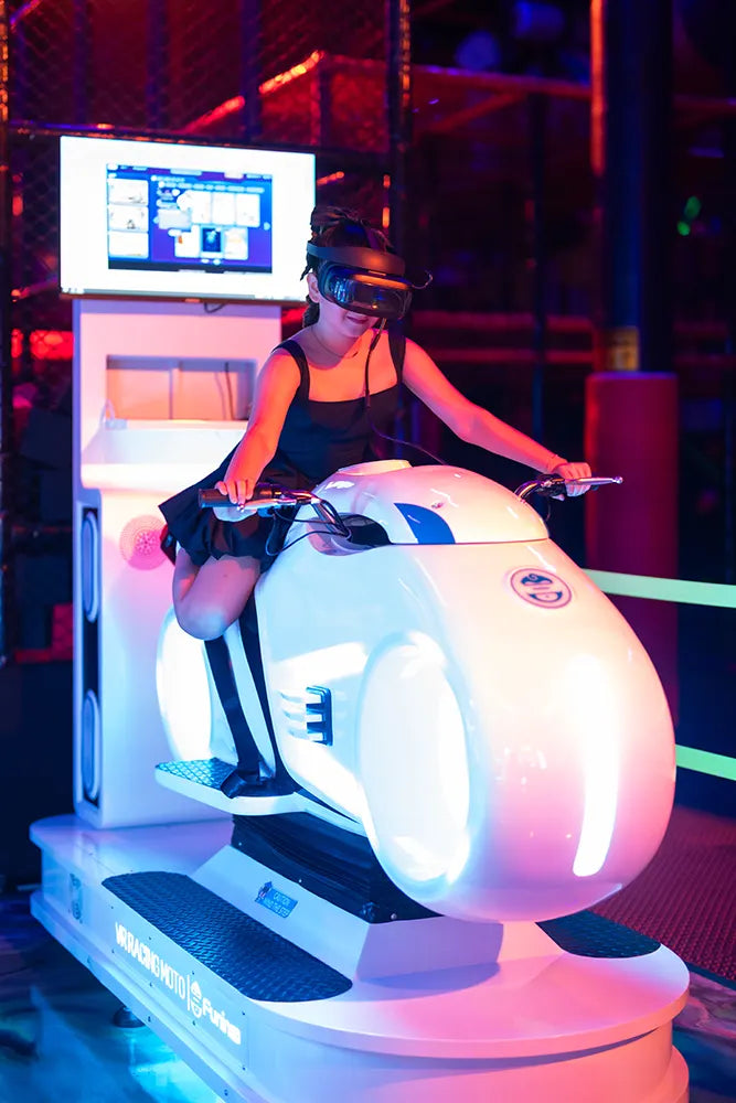 A person wearing a VR headset sits on a glowing white motorcycle simulator, gripping the handlebars, in an arcade with colorful neon lights. A screen in front displays the virtual experience.