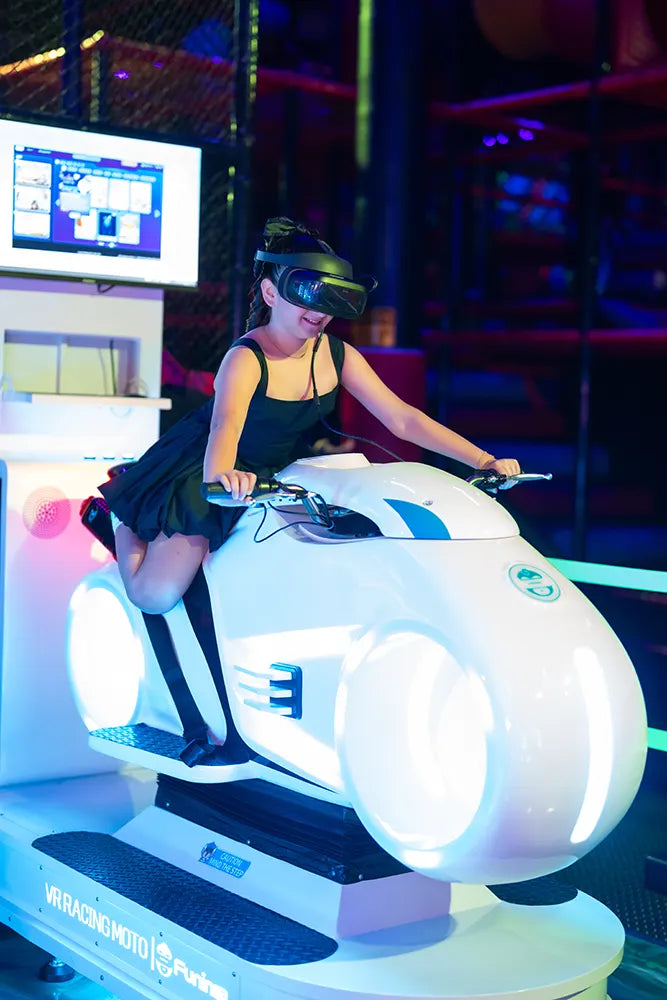A woman wearing a black dress and VR headset rides a futuristic-style motorcycle simulator in an indoor arcade with colorful lighting.