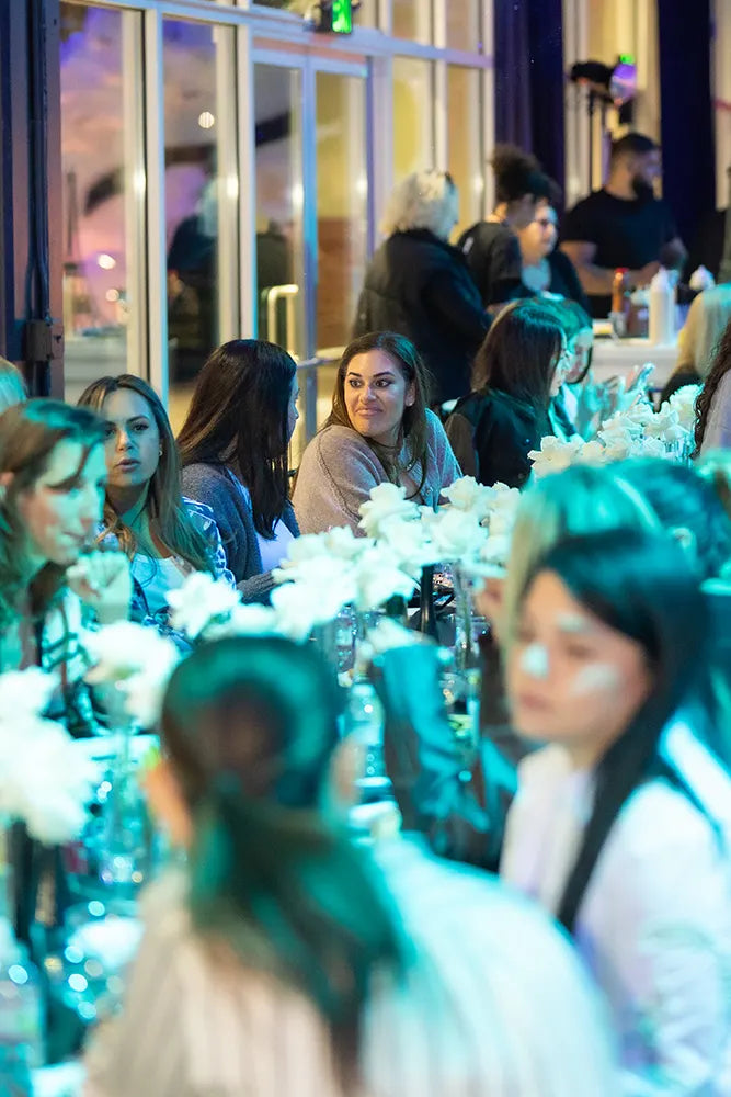 A group of people sit at a long, flower-decorated table indoors, engaged in conversation. The atmosphere is lively, and the lighting is bright and colorful.