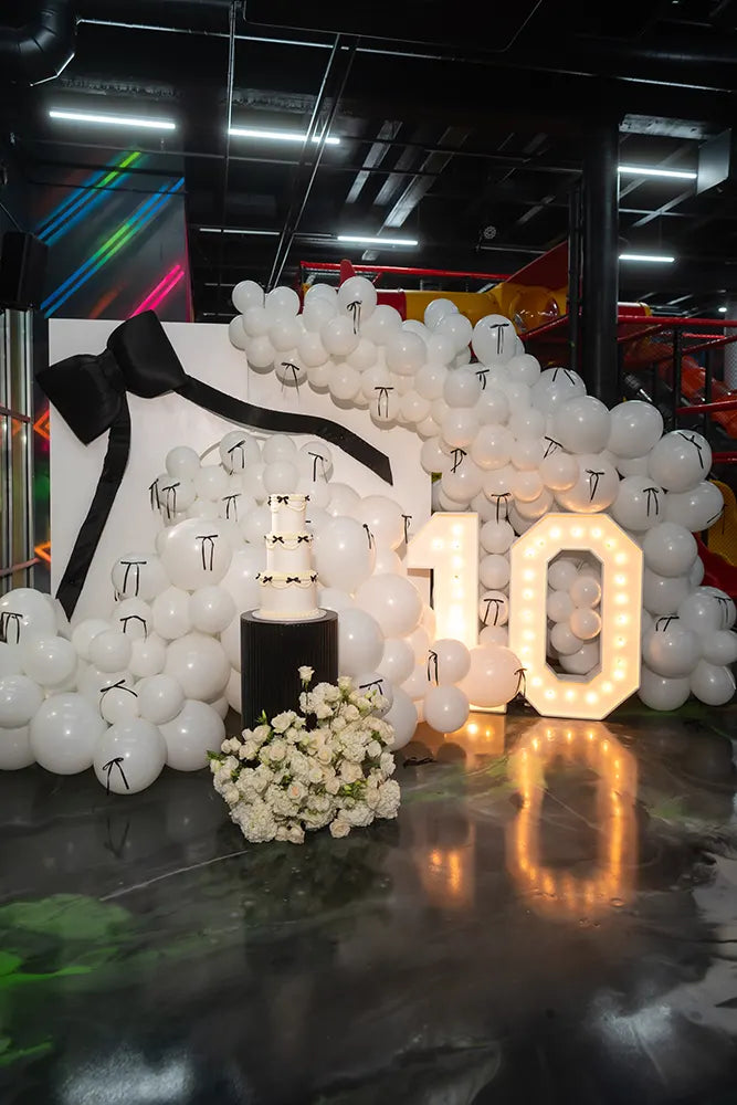 A festive display featuring a large cluster of white balloons, a white cube with a black ribbon, a tiered cake, a bouquet of white flowers, and illuminated numbers 