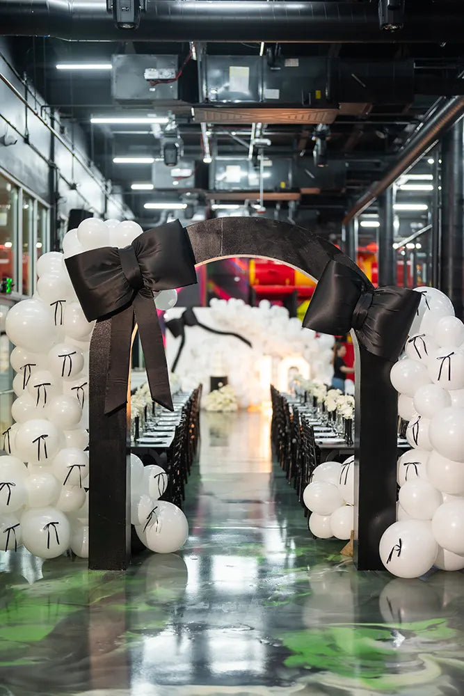A black arch decorated with large black bows stands at the entrance to a long dining table, flanked by white balloons, in a modern, brightly lit indoor event space.