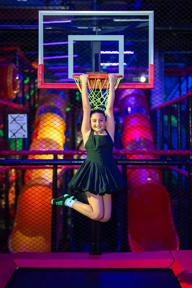 A young girl in a black dress hangs from a basketball hoop, smiling, at an indoor play center with colorful slides and bright lights in the background.