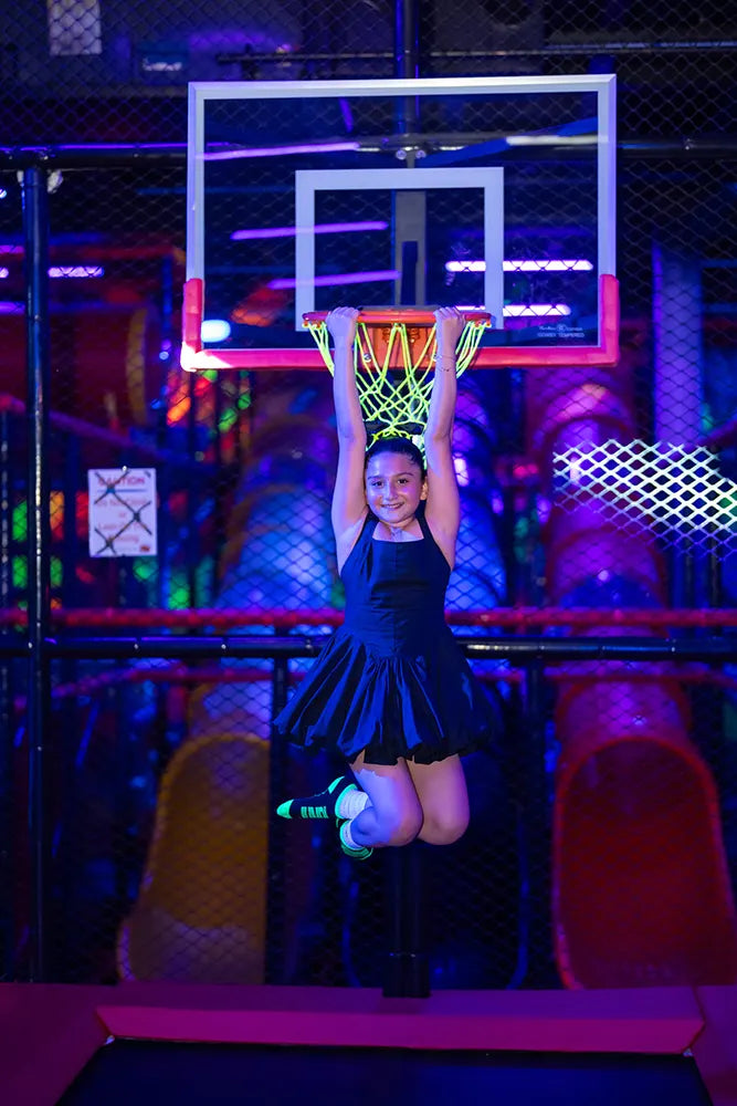 A young girl in a black dress hangs from a neon-lit basketball hoop with both hands, smiling, in a colorful indoor play area with glowing lights and vibrant colors in the background.