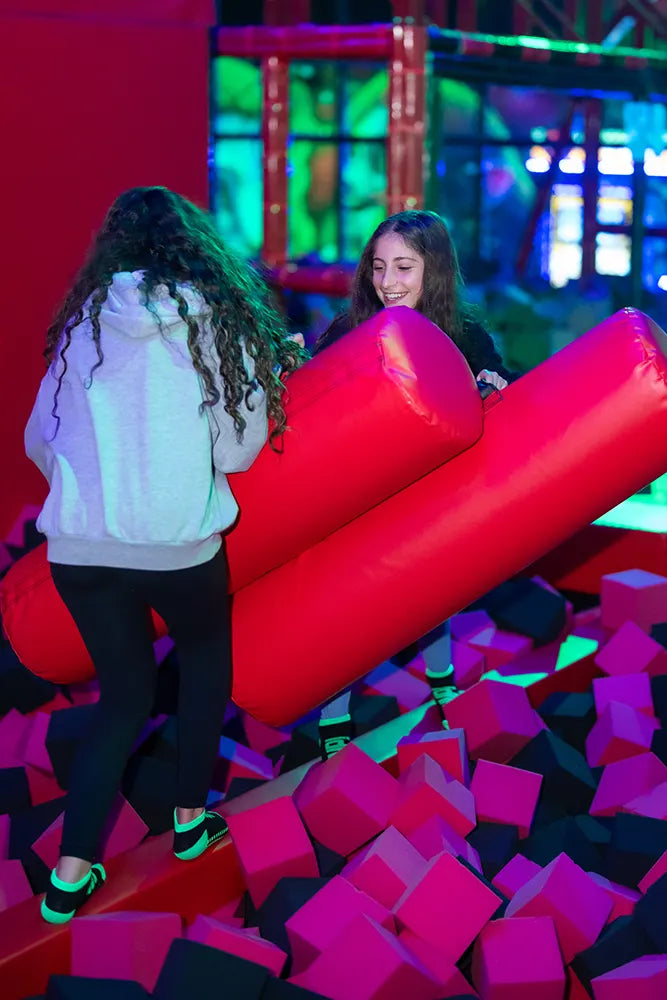 Two girls stand in a foam pit, playfully jousting with large red padded sticks at an indoor activity center with colorful lighting and foam blocks around them.