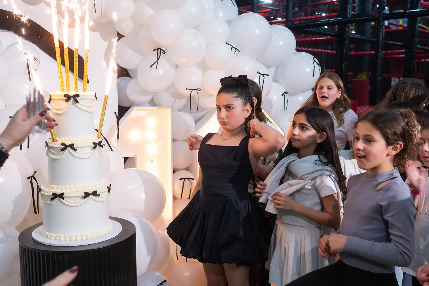 A girl in a black dress stands with friends in front of a tall white birthday cake with sparklers, surrounded by white balloons at a party. She looks serious while others watch the cake being presented.