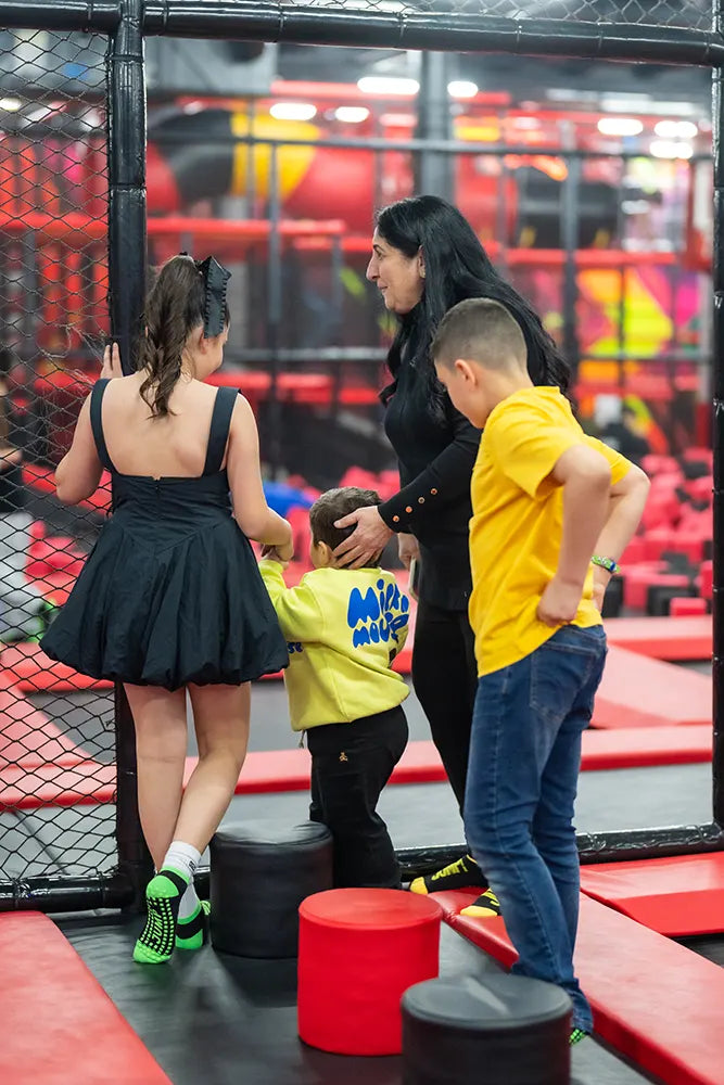 A woman and three children stand together inside an indoor play area with red and black padded obstacles and nets. The children appear to be chatting or preparing to play.