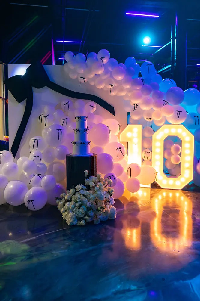 A decorated event space with a large arrangement of white balloons, a big black bow, a tiered cake, a bouquet of white flowers, and an illuminated “10” sign, all under colorful blue and purple lighting.