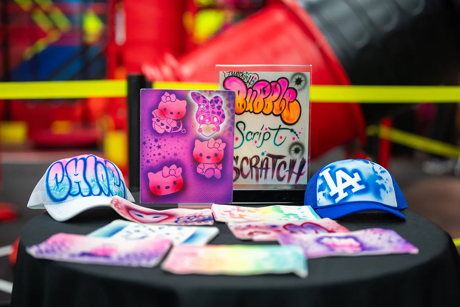 A display of colorful airbrushed art, including hats, canvas art featuring Hello Kitty, and signs with vibrant graffiti-style lettering, arranged on a round black table with a blurred playground background.