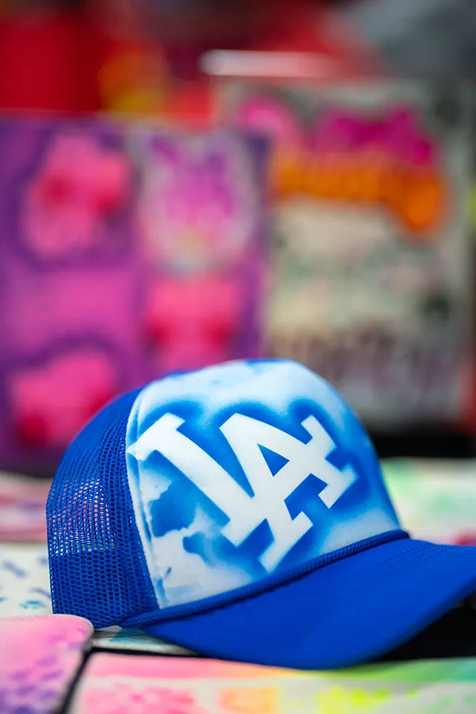 A blue and white mesh trucker hat with “LA” spray-painted on the front is placed on a colorful, graffiti-style background. The hat is in sharp focus, while the background is blurred.