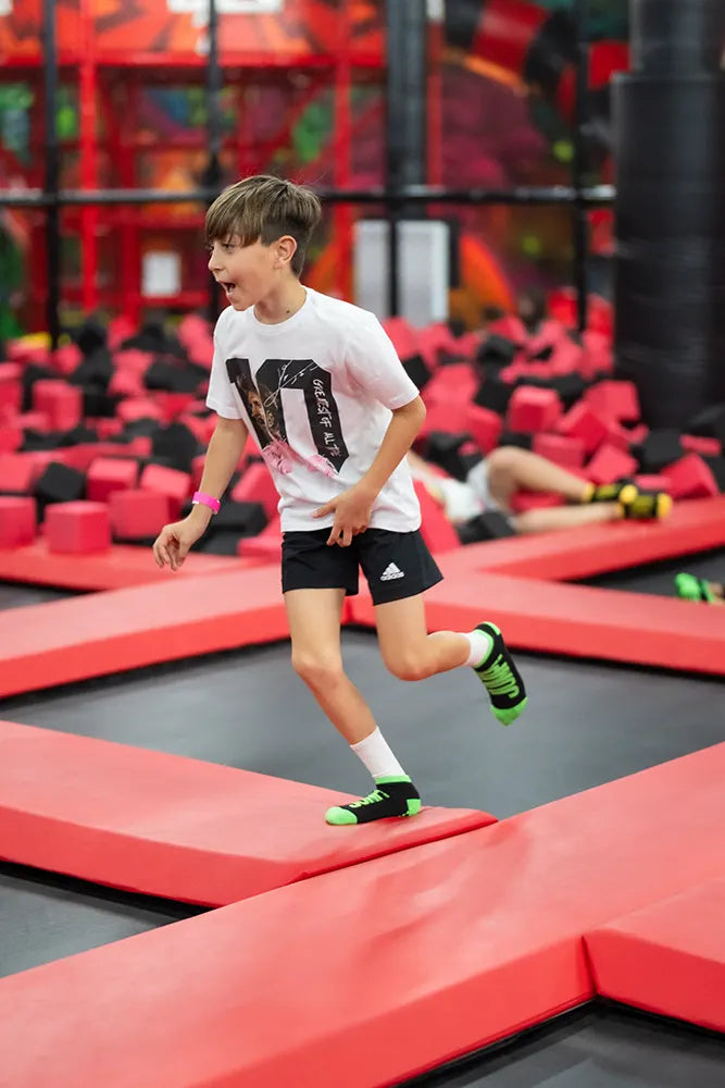A young boy in a white T-shirt, black shorts, and green socks runs on red padded surfaces at an indoor trampoline park. Foam cubes and another child are visible in the background.