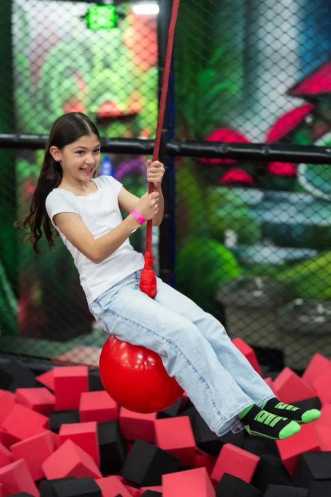 A smiling girl in a white T-shirt and jeans swings on a red rope ball above a foam pit filled with red and black blocks at an indoor play center.