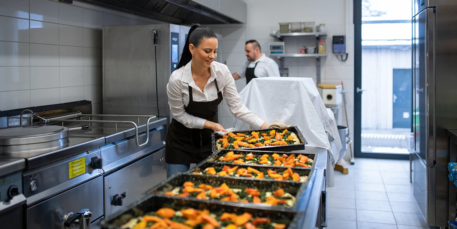 A woman in an apron places a tray of food with vegetables on a counter in a commercial kitchen, while a man works in the background. Several trays of prepared food are lined up in front of her.
