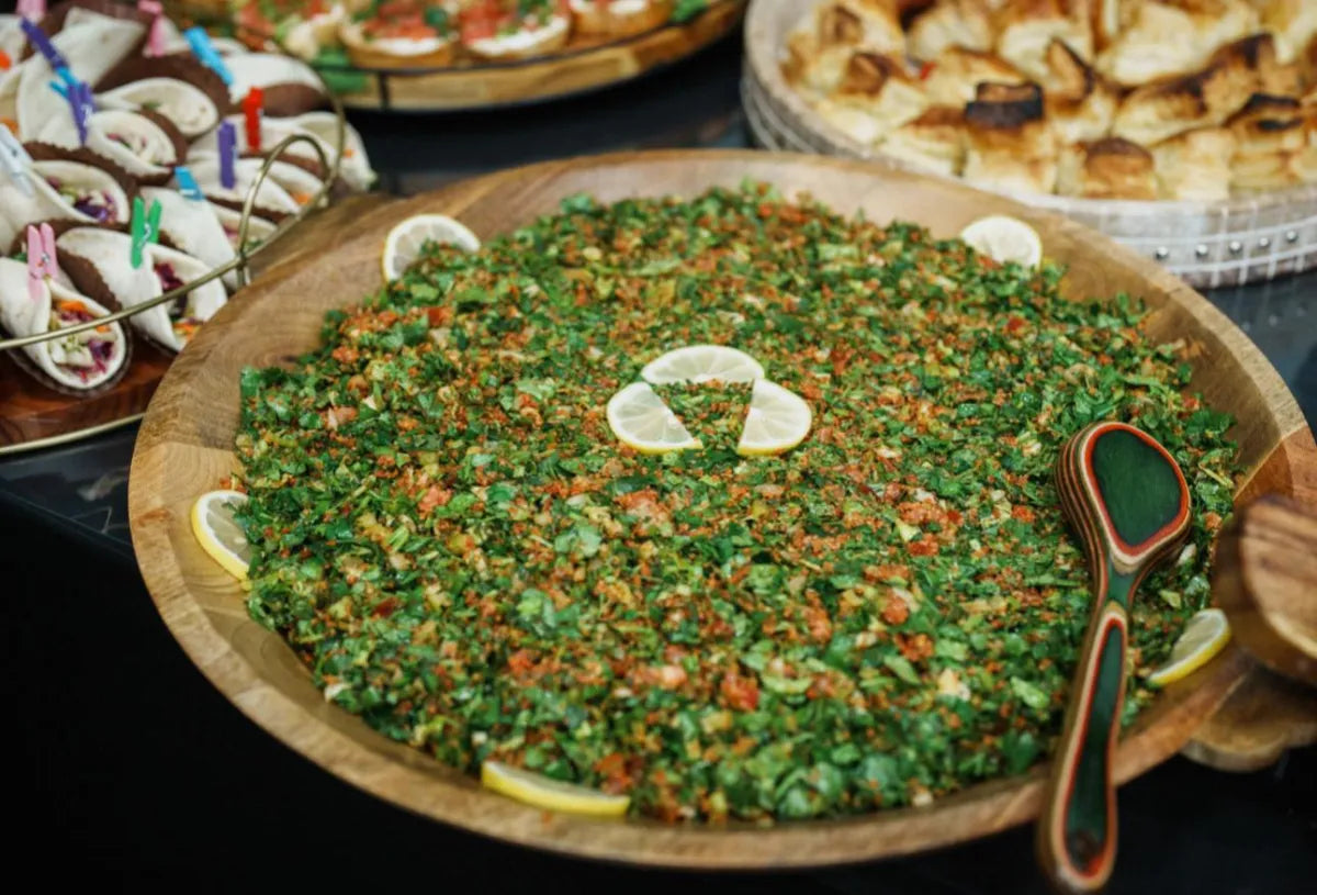 A large wooden platter filled with tabbouleh salad, garnished with lemon slices around the edges. A wooden serving spoon rests on the right. Other assorted dishes are visible in the background.