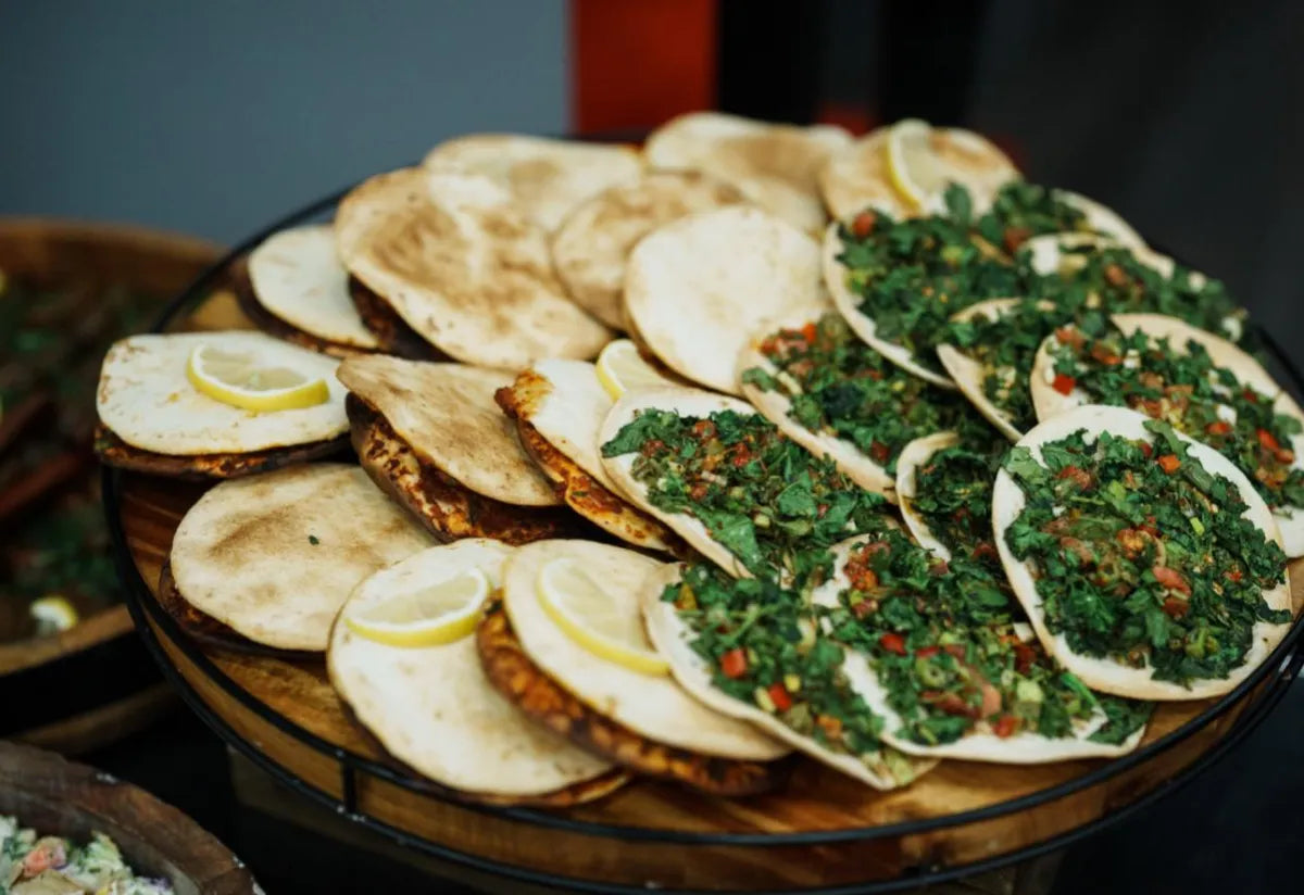 A platter of Middle Eastern flatbreads, some topped with chopped greens, tomatoes, and herbs, and others plain, garnished with lemon wedges.
