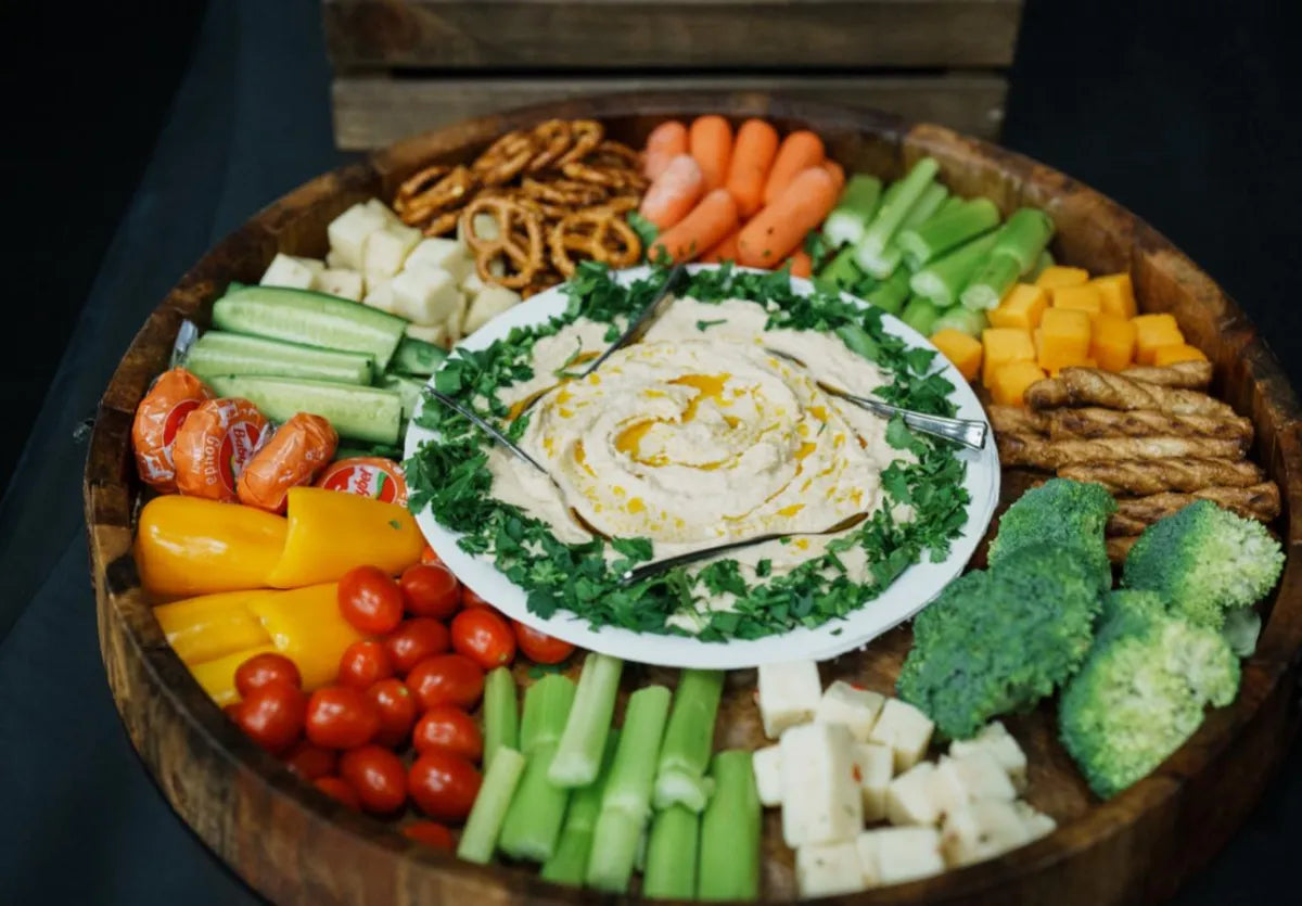 A round wooden tray holds a plate of hummus topped with oil and herbs, surrounded by assorted fresh vegetables, cheese cubes, cherry tomatoes, pretzels, and breadsticks arranged in sections.