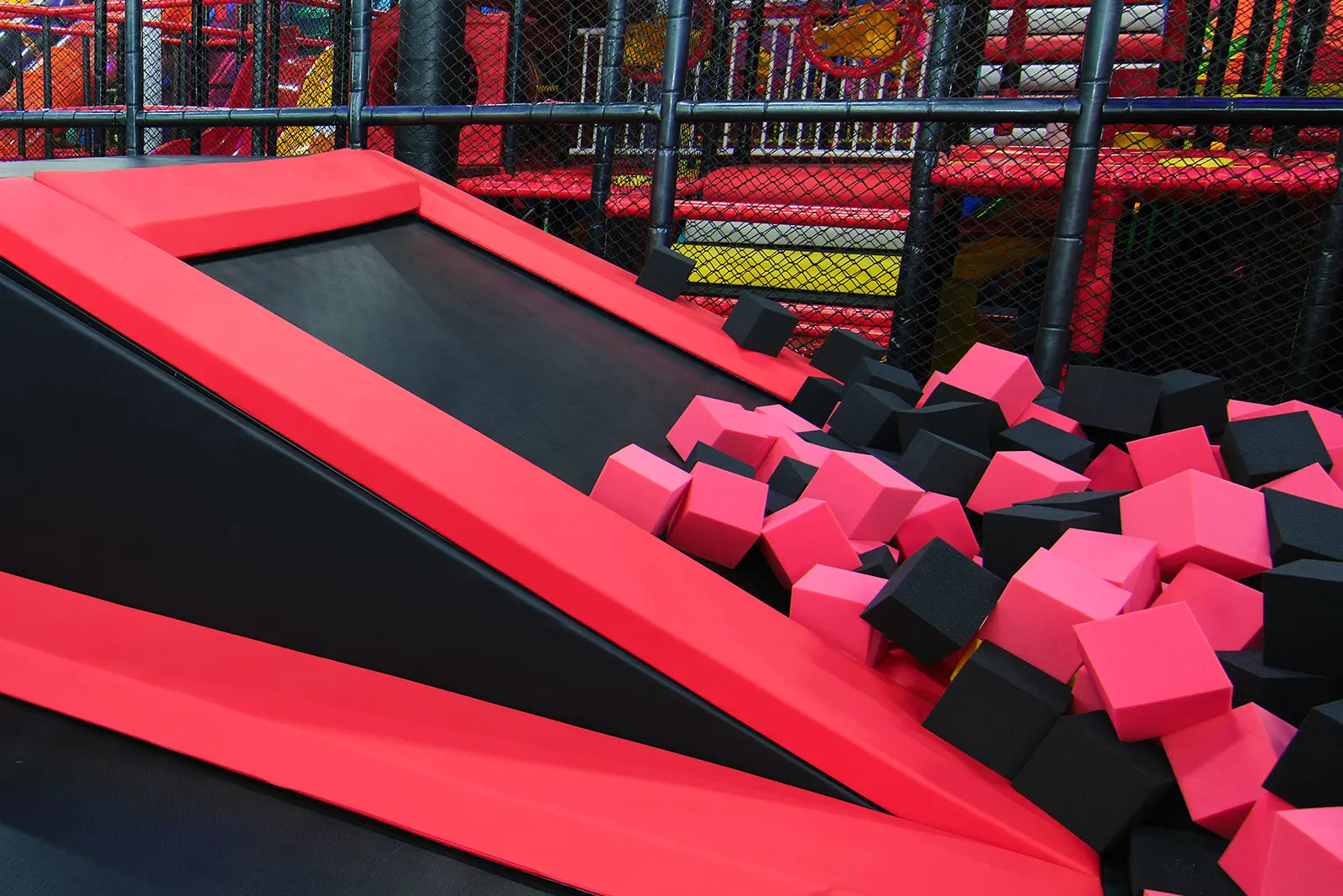 A trampoline park area with a black and red sloped trampoline leading into a foam pit filled with pink and black foam cubes. Bright play structures and safety netting are visible in the background.
