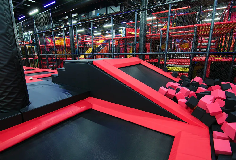 Indoor trampoline park featuring black trampolines with red padding, a foam pit filled with pink and black cubes, netted barriers, and colorful play structures in the background.