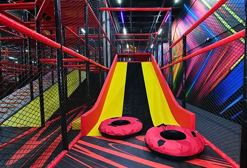 A brightly colored indoor slide with yellow and black lanes, red railings, netted sides, and two red inflatable tubes at the bottom, set in a vibrant play area with colorful wall designs.