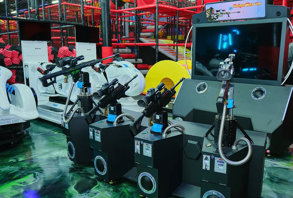 Three black arcade shooting game machines with mounted guns are positioned in front of a large screen display, with white virtual reality chairs and a colorful indoor play area in the background.