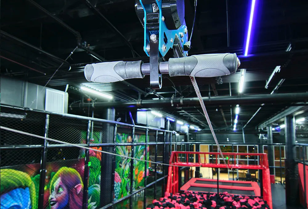 A close-up of a zip line handle inside an indoor adventure park, with a foam pit below, safety netting, and colorful wall art featuring fantasy characters in the background. Bright lights illuminate the area.