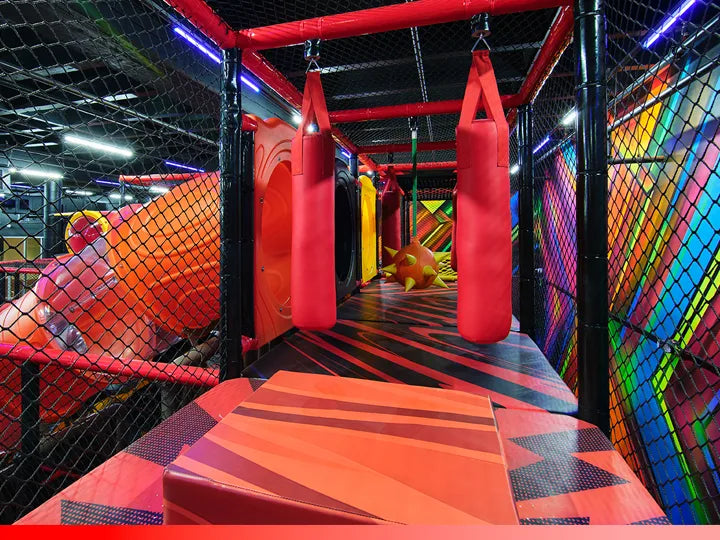 Indoor playground with red hanging punching bags, padded platforms, a netted enclosure, and colorful geometric walls and flooring. A yellow climbing structure and enclosed slide are visible in the background.