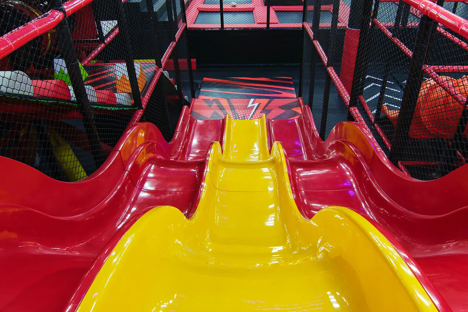 A brightly colored indoor playground slide with three lanes: two red on the sides and one yellow in the center, surrounded by safety netting and red railings. The slide leads down to a black and red floor.