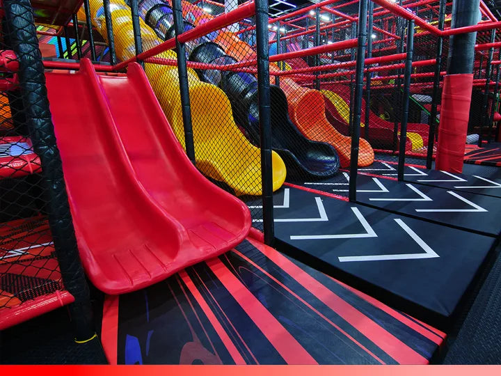 A vibrant indoor playground features red, yellow, and black slides, padded flooring with white arrows, and climbing structures enclosed by safety netting.