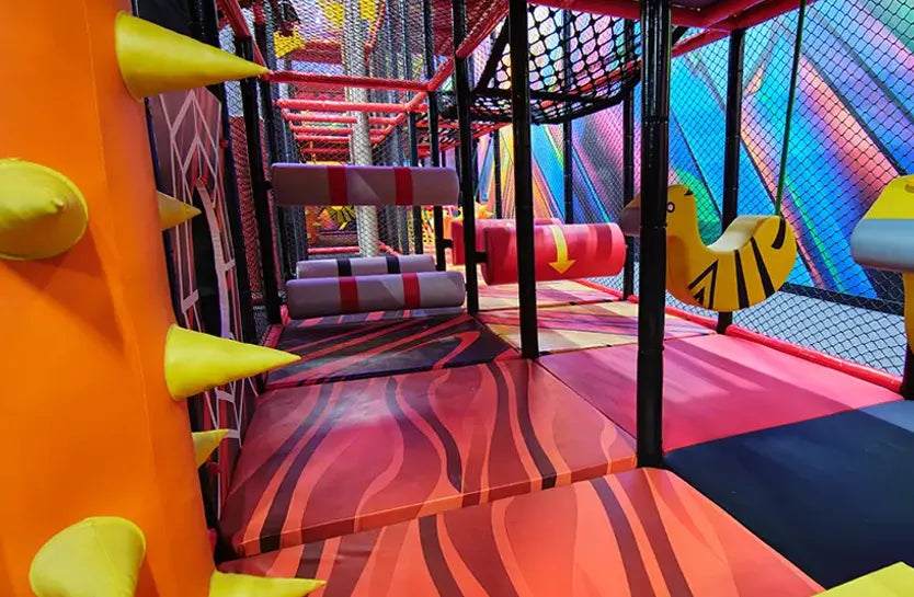 Indoor playground with colorful padded flooring, foam obstacles shaped like spikes and logs, animal-shaped swings, and safety netting, designed for children’s climbing and play activities.