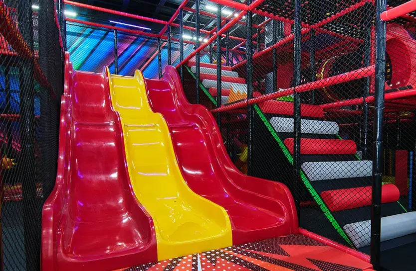 A brightly colored indoor playground features two red slides and a yellow slide, surrounded by black safety nets and padded climbing areas with red, gray, and green steps.