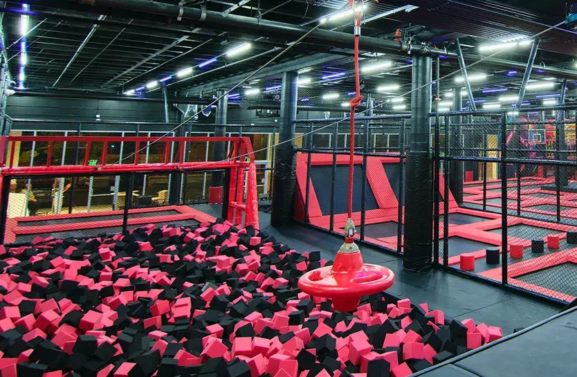 Indoor trampoline park with black and red foam pits, trampoline areas, a zipline with a red seat, and safety nets, all brightly lit with overhead lighting.