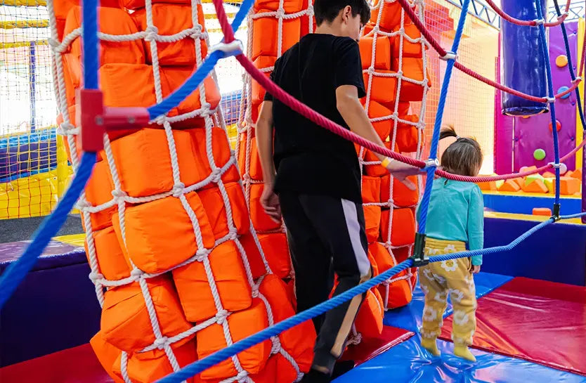 Two children, one older and one younger, walk through a colorful indoor playground. They are surrounded by orange padded climbing structures and white ropes. The area has bright mats and netting, creating a fun and safe environment.