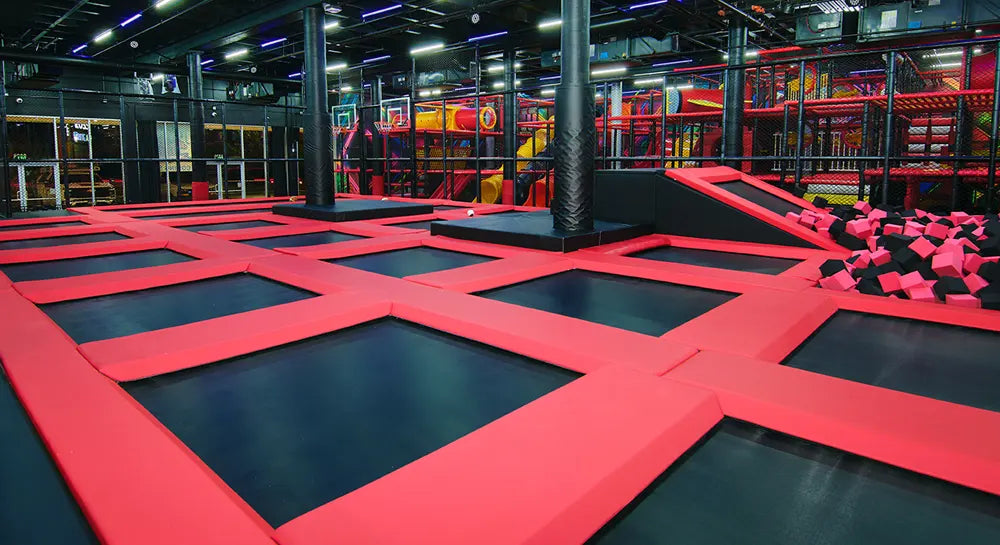 An indoor trampoline park with interconnected red and black trampolines, foam pit with black and pink cubes, and colorful climbing structures in the background under bright overhead lights.