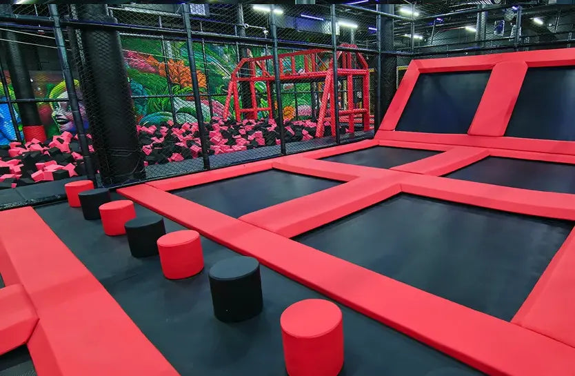 Indoor trampoline park with black and red trampolines, foam pits, padded stepping stones, and climbing structures, set against a colorful wall mural. The area is enclosed with safety netting.